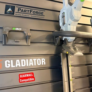 May include: A gray wall-mounted storage system with the text "PartForge" and "Gladiator". An EGO tool holder is mounted on the wall. Two green and gray EGO tools are also mounted on the wall. The wall is compatible with Gearwall.