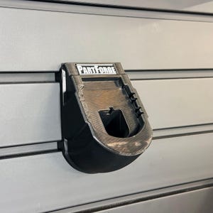 May include: A black plastic tool mount featuring the text PartForge printed in white on the top edge. The mount is designed with a recessed center and a curved lower section, securely attached to a horizontal gray slat wall panel.