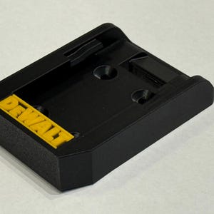 May include: Black plastic power tool battery adapter with the yellow text "DEWALT". The adapter has a rectangular shape with rounded edges and two holes. It is designed to connect to a power tool.