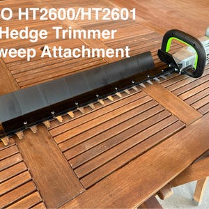 Peut inclure: Une tondeuse de haie EGO HT2600/HT2601 avec un accessoire de balayage. La tondeuse est grise et noire avec un accent vert. L'accessoire de balayage est noir et comporte une rangée de dents. Le texte "EGO HT2600/HT2601 Hedge Trimmer Sweep Attachment" est visible.