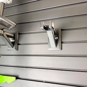 May include: A close-up of a gray wall with mounted tool holders. The holders are made of metal and are designed to hold gardening tools. The wall is made of horizontal panels. The tools are partially visible.