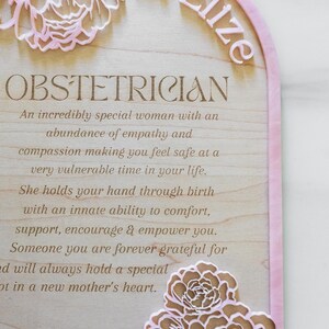 Puede incluir: Una placa de madera arqueada con un borde de acr&iacute;lico rosa que presenta el texto Dr Elize, OBSTETRICIAN, y un mensaje sobre empat&iacute;a y apoyo durante el parto. El dise&ntilde;o incluye flores rosas cortadas con l&aacute;ser y los nombres Courtney, Josh y baby Amelia.