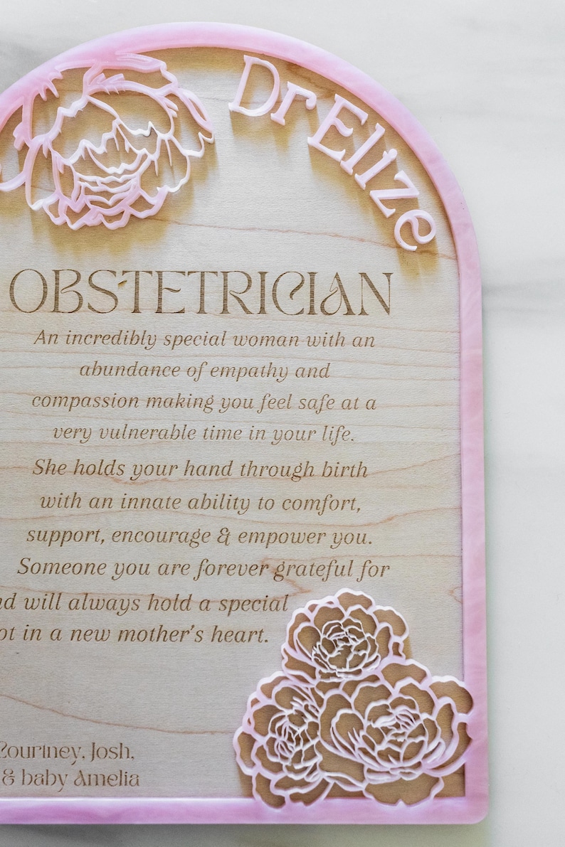 Puede incluir: Una placa de madera arqueada con un borde de acr&iacute;lico rosa que presenta el texto Dr Elize, OBSTETRICIAN, y un mensaje sobre empat&iacute;a y apoyo durante el parto. El dise&ntilde;o incluye flores rosas cortadas con l&aacute;ser y los nombres Courtney, Josh y baby Amelia.