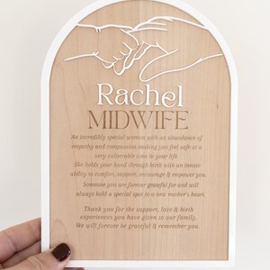 Puede incluir: Una placa de madera arqueada con borde blanco que presenta un recorte blanco de dos manos entrelazadas. El texto dice: Rachel MIDWIFE, seguido de un p&aacute;rrafo que expresa gratitud por el apoyo, la empat&iacute;a y la compasi&oacute;n recibidos.