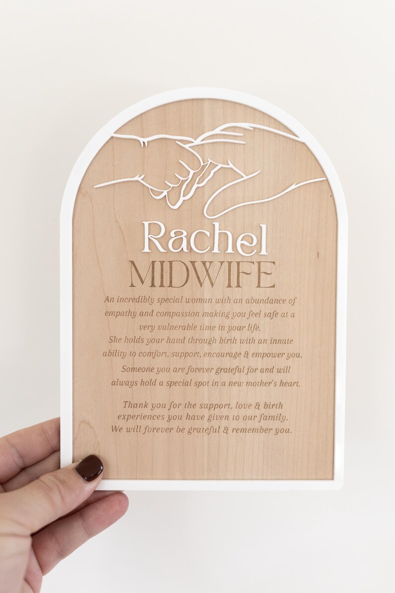 Puede incluir: Una placa de madera arqueada con borde blanco que presenta un recorte blanco de dos manos entrelazadas. El texto dice: Rachel MIDWIFE, seguido de un p&aacute;rrafo que expresa gratitud por el apoyo, la empat&iacute;a y la compasi&oacute;n recibidos.