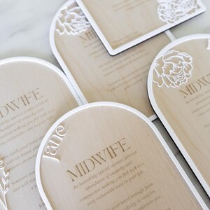 Puede incluir: Placas de madera en forma de arco con bordes blancos y recortes florales blancos. Cada placa presenta el texto MIDWIFE seguido de un poema sobre empat&iacute;a y apoyo. Una placa incluye el nombre Jane en letras cursivas blancas.