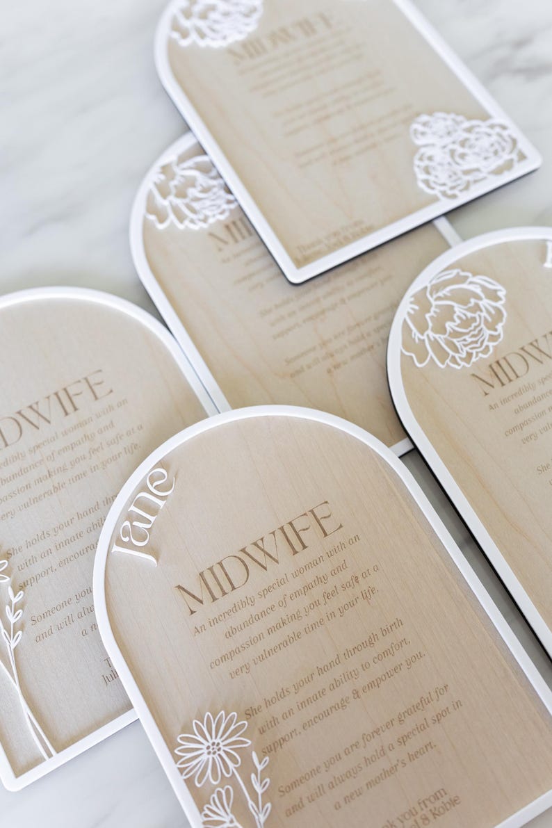 Puede incluir: Placas de madera en forma de arco con bordes blancos y recortes florales blancos. Cada placa presenta el texto MIDWIFE seguido de un poema sobre empat&iacute;a y apoyo. Una placa incluye el nombre Jane en letras cursivas blancas.