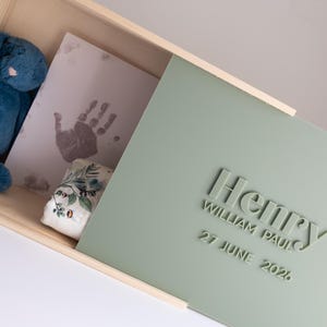 May include: A light wood keepsake box with a sliding sage green lid featuring the raised text Henry, William Paul, and 27 June 2026. Inside the box sits a blue plush rabbit, a white card with a grey handprint, and a small floral patterned item.
