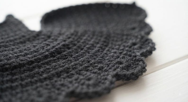 May include: A dark charcoal gray crocheted textile piece featuring a dense, textured stitch pattern throughout the main body and a decorative, scalloped edge finish along the curved border, showcasing the intricate yarn loops and construction.