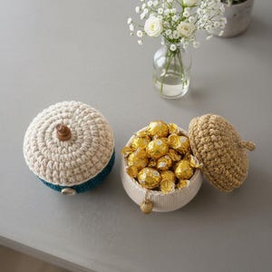 Puede incluir: Recipiente de dulces en forma de bellota tejido a ganchillo con tapa, lleno de bombones envueltos en oro. El recipiente es de color beige y verde azulado, con un cierre de botón de madera. Un pequeño jarrón de flores blancas y una suculenta en maceta están en el fondo.