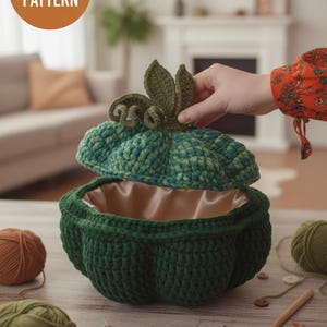 May include: A handmade crochet pumpkin-shaped basket with a lid. The basket is green with a teal and green lid and a satin interior. The lid is being lifted to reveal the inside. The image also includes the text "CROCHET PATTERN".