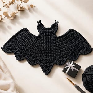 May include: A black crocheted bat applique featuring a textured stitch pattern with pointed ears and scalloped wing edges. The bat is crafted from dark yarn and displays a symmetrical shape with a central body and wide, outstretched wings.