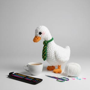 Può includere: Un'oca bianca all'uncinetto con una cravatta a righe verdi e gialle, con zampe e becco arancioni. Sono presenti anche una tazza bianca, un astuccio nero con uncinetti, forbici e un gomitolo di filo bianco.
