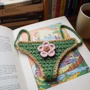 Op de afbeelding: Een handgemaakt groen gehaakt bladwijzer in de vorm van ondergoed, met een roze bloem en gouden rand. De bladwijzer ligt op een open boek met een kleurrijke illustratie. De bladwijzer heeft groene bandjes.