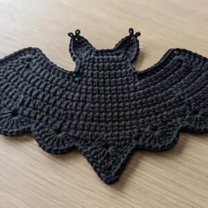 May include: A black crochet bat applique featuring a textured stitch pattern with scalloped wing edges and two pointed ears. The bat is crafted from dark yarn and displays a symmetrical shape with a central body and wide, outstretched wings.