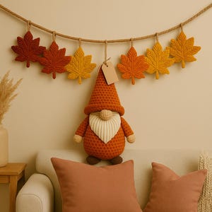 May include: A seasonal display featuring a crochet gnome with an orange hat and beard, and a garland of maple leaf ornaments in shades of orange and yellow. The gnome is positioned below the garland, creating a cosy autumn scene.