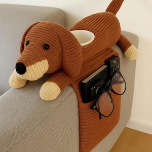 Può includere: Un organizer per bracciolo del divano a forma di bassotto lavorato a maglia marrone. Il cane ha pancia, zampe e muso bianchi. È dotato di un portabicchieri, una tasca con telecomando e telefono e un paio di occhiali neri.