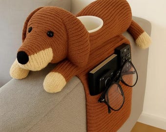 May include: A brown knitted dachshund-shaped sofa arm organizer. The dog has a white belly, paws, and snout. It features a cup holder, a pocket with a remote and a phone, and a pair of black glasses.