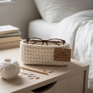 May include: A cream-colored, crocheted glasses holder with a pair of eyeglasses inside. A tag reads "Comfy Glasses Holder - Beginner Crochet Pattern." A ball of yarn, scissors, and crochet hooks sit nearby on a wooden nightstand.