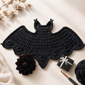 May include: A black crocheted bat applique featuring a textured stitch pattern with scalloped wing edges. The bat is positioned alongside a dark purple dahlia flower, a small black gift box with a white ribbon, a metal crochet hook, and yarn.