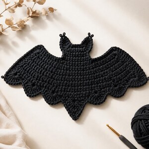 May include: A black crochet bat applique featuring a central body with two pointed ears and wide, scalloped wings. The piece is crafted with a dense, textured stitch pattern, creating a flat, symmetrical silhouette of a bat in flight.