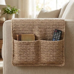 May include: A crocheted, tan-colored sofa arm organizer with two pockets. One pocket holds a small book, and the other holds a black remote control. The organizer is designed to fit over the arm of a sofa.