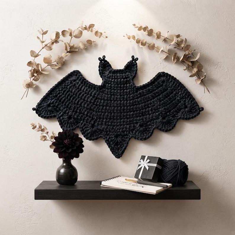 May include: A black crocheted bat wall hanging with scalloped wing edges and pointed ears. The bat is displayed above a black shelf holding a dark flower in a small vase, a black gift box with a white ribbon, a ball of black yarn, and a notebook.