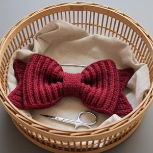 Könnte beinhalten: Eine weinrote, gestrickte Fliege liegt in einem runden Weidenkorb, der mit beigem Stoff ausgekleidet ist. Eine kleine, silberne Schere befindet sich ebenfalls im Korb. Die Fliege hat eine gerippte Textur.