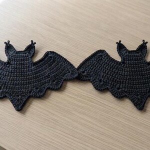 May include: Two identical black crochet bat appliques featuring detailed wing structures, pointed ears, and scalloped edges. Each piece is crafted with a dense yarn stitch pattern, creating a flat, symmetrical silhouette of a bat with outstretched wings.