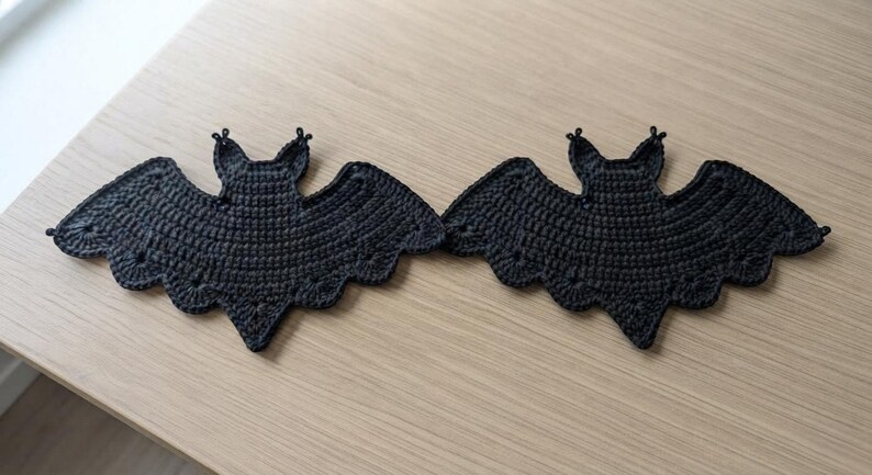 May include: Two identical black crochet bat appliques featuring detailed wing structures, pointed ears, and scalloped edges. Each piece is crafted with a dense yarn stitch pattern, creating a flat, symmetrical silhouette of a bat with outstretched wings.