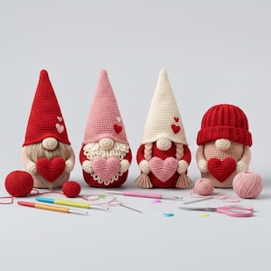 Puede incluir: Cuatro gnomos de ganchillo hechos a mano en colores rojo, rosa y crema, cada uno sosteniendo un corazón. Los gnomos tienen sombreros largos y puntiagudos y barbas blancas. Agujas de ganchillo, hilo y tijeras están dispuestas frente a los gnomos, sugiriendo un tema artesanal.