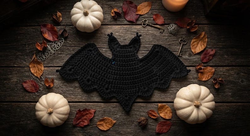 May include: A black crocheted bat ornament with scalloped wing edges. The bat is surrounded by two white pumpkins, scattered dried autumn leaves, and two small metal keys on a dark, weathered wooden surface with a glowing candle nearby.