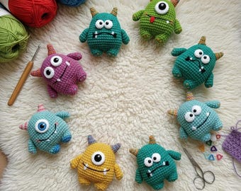 May include: A collection of handmade crochet monster toys in various colors, including green, blue, purple, and yellow. Each monster has large eyes, horns, and a unique expression. The toys are arranged on a white, fluffy surface, with yarn and crochet tools visible.