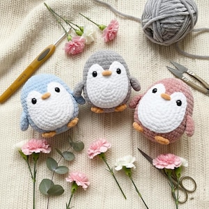 May include: Three crocheted penguin toys in blue, grey, and pink, each with a white belly, black eyes, and orange beak and feet. A crochet hook, yarn, and scissors are also present, along with pink and white carnation flowers.