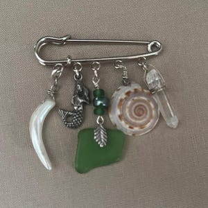 May include: A silver safety pin brooch with dangling charms. Charms include a white curved tooth, a mermaid, green beads, a seashell, a piece of green sea glass, and a clear crystal point. The brooch is on a neutral background.