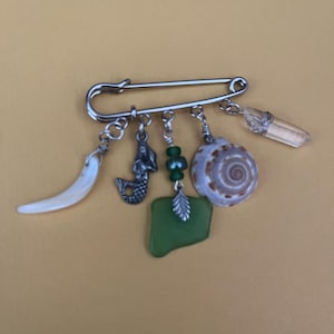 May include: A silver safety pin brooch with dangling charms. Charms include a white tusk, a mermaid, green beads, a green sea glass piece, a seashell, and a clear crystal point. The brooch is set against a yellow background.