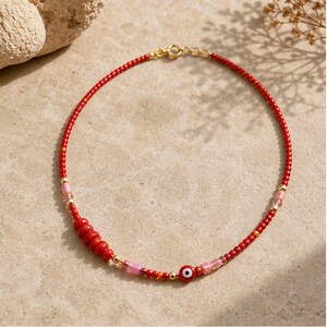 May include: A beaded necklace featuring small red seed beads, interspersed with metallic gold spheres, translucent pink cylindrical beads, and a single red evil eye charm. The necklace is secured with a gold-toned metal lobster claw clasp.