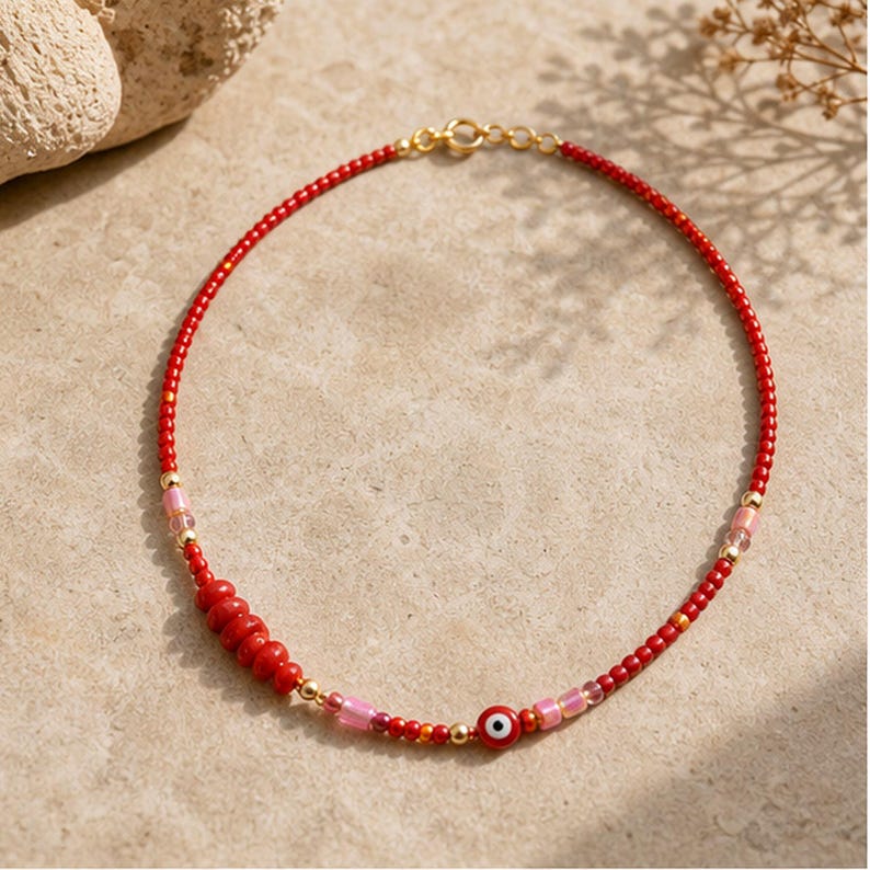 May include: A beaded necklace featuring small red seed beads, interspersed with metallic gold spheres, translucent pink cylindrical beads, and a single red evil eye charm. The necklace is secured with a gold-toned metal lobster claw clasp.
