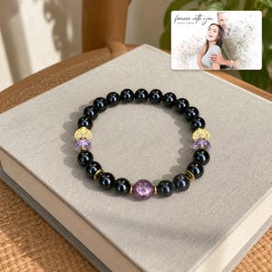 May include: A beaded bracelet featuring smooth black spherical beads, three translucent purple faceted beads, and four gold-toned textured metal spacers. A small rectangular card is visible with the text "forever with you" and "OLIVIA & ADAM" above a photo of a couple.