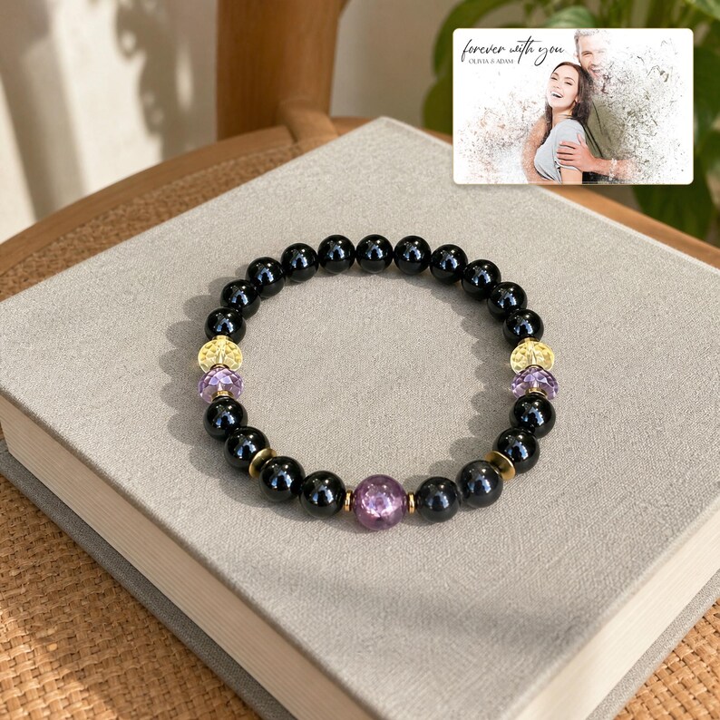 May include: A beaded bracelet featuring smooth black spherical beads, three translucent purple faceted beads, and four gold-toned textured metal spacers. A small rectangular card is visible with the text "forever with you" and "OLIVIA & ADAM" above a photo of a couple.