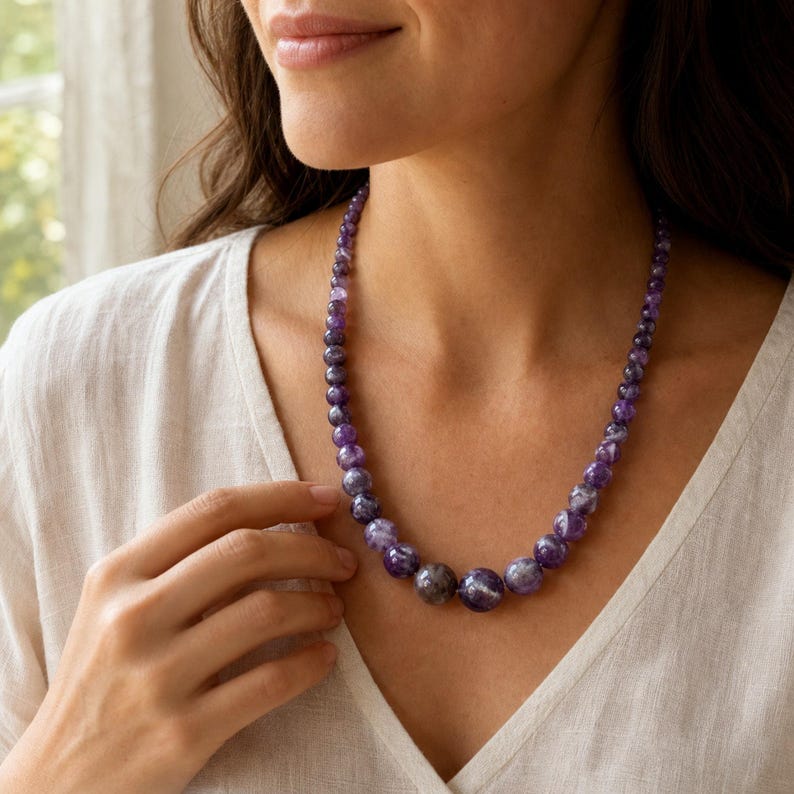 May include: A beaded necklace featuring round, polished amethyst stones in varying shades of deep purple and violet. The beads are graduated in size, with the largest stones positioned at the center and smaller beads tapering toward the back.