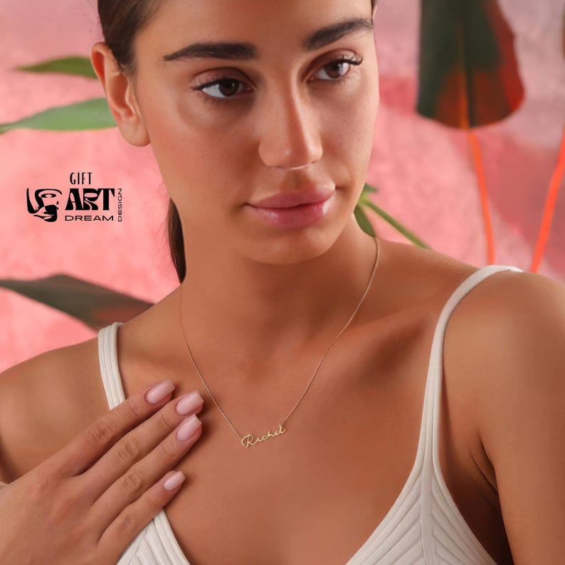 May include: A delicate gold necklace with the name "Rachel" in cursive script. The necklace is worn by a person wearing a white top. The background is a soft pink and white abstract design. The logo "GIFT ART DREAM DESIGN" is visible in the upper left corner.