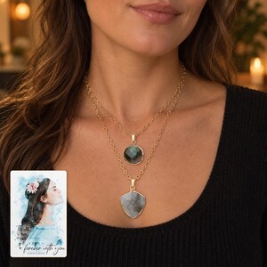 Grey-green round and shield-shaped pendants on a layered gold-tone necklace, beside a personalized digital greeting card.