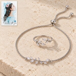 Silver-tone leaf bracelet and matching open-vine ring arranged on ivory travertine, showing clear marquise accents and a sliding box-chain closure. A personalized digital greeting card is shown alongside.