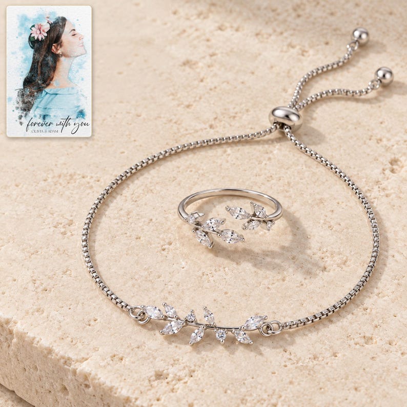 Silver-tone leaf bracelet and matching open-vine ring arranged on ivory travertine, showing clear marquise accents and a sliding box-chain closure. A personalized digital greeting card is shown alongside.