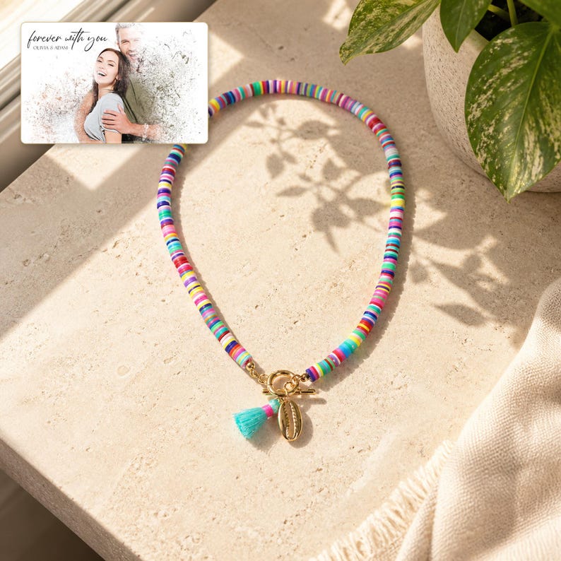 May include: A necklace made of small, multicolored clay disc beads featuring a gold-toned toggle clasp, a small turquoise tassel, and a gold-toned cowrie shell charm. A rectangular card displays a photo of a couple with the text "forever with you".