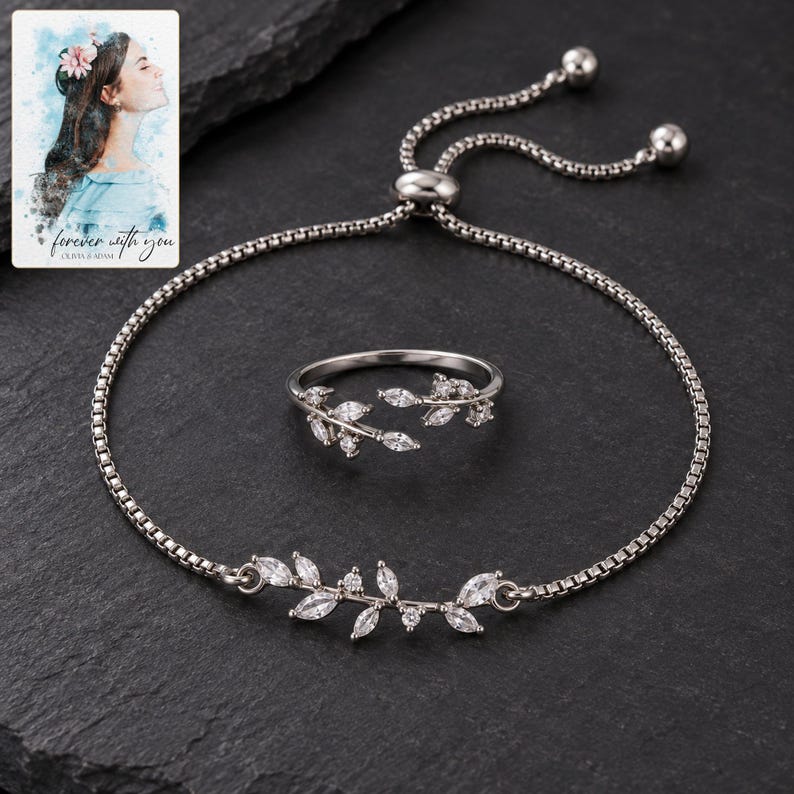 Clear leaf-accent bracelet and matching ring on charcoal slate, showing the polished slider and two ball-ended chain tails. A personalized digital greeting card is shown alongside.