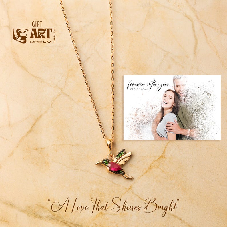 May include: A gold-colored hummingbird pendant necklace with a red gemstone and green accents. The necklace hangs on a gold chain. A card with the text "forever with you" and a couple's photo is in the background. The phrase "A Love That Shines Bright" is at the bottom.