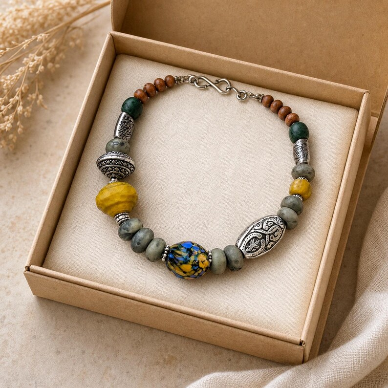 Puede incluir: Una pulsera de cuentas con una mezcla de cuentas de madera marr&oacute;n, cuentas de piedra gris, cuentas amarillas texturizadas y una cuenta central de vidrio moteado en azul y amarillo. Incluye espaciadores de metal plateado y un cierre de gancho.