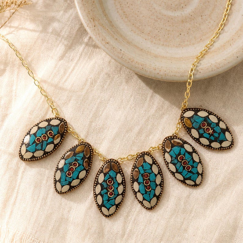 May include: A gold-tone chain necklace featuring five oval-shaped pendants. Each pendant displays a mosaic pattern of turquoise blue, cream, and dark brown segments, accented with small, round, reddish-brown stones arranged in a vertical line.